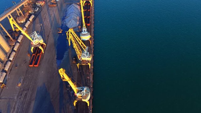 Yellow cargo cranes at a sea dock. Aerial view of heavy industrial machinery standing in a row next to deep blue water with long shadows.