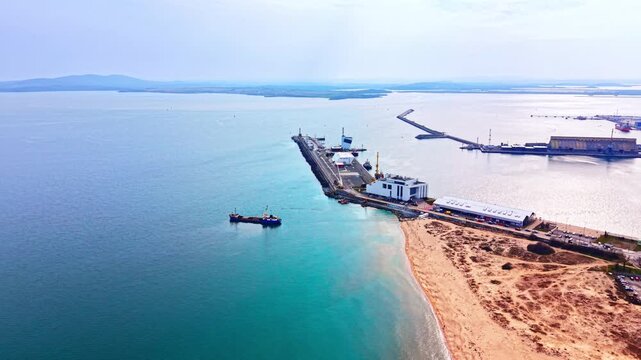 Coastal breakwater pier and sandy beach area, Burgas, Bulgaria. Broad aerial drone view of a maritime jetty, a building, a sandy shore and turquoise sea water under an overcast sky.