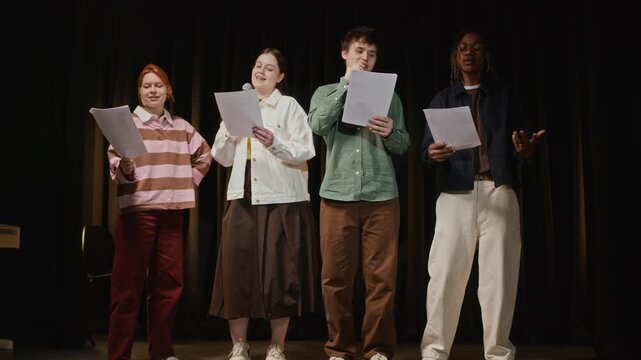 Full length shot of diverse teenagers standing on stage, holding scripts and rehearsing musical performance with microphones. For school drama club promotions and arts education marketing campaigns