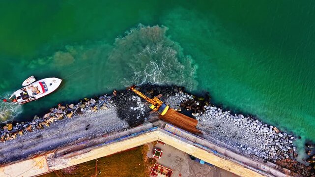 Aerial view of harbor breakwater construction with excavator and boat. Yellow excavator on rocky jetty, white vessel moored alongside turquoise water.