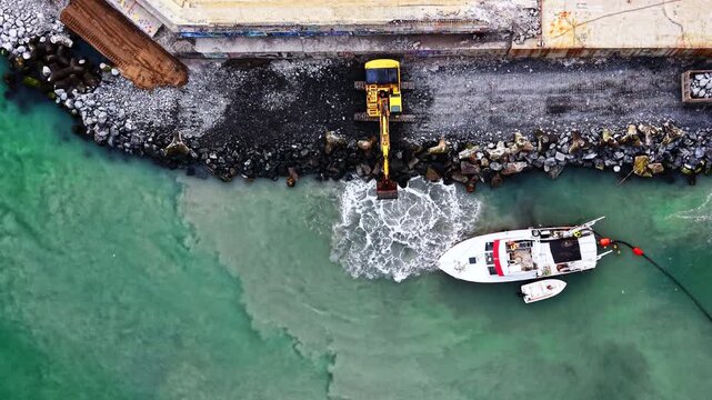 Overhead drone perspective of dredging vessel and excavator at port breakwater. Yellow excavator digs into rocky seawall as white boat floats on turquoise water below.