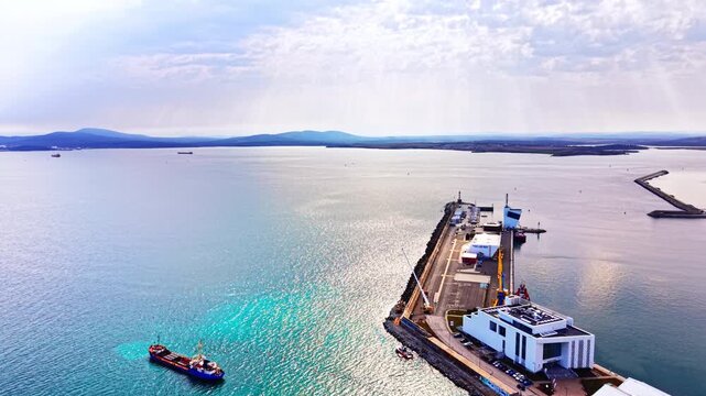 Sea port pier with cargo ship and mountain backdrop, Bulgaria. Drone shot of narrow quay extending into calm sea, white terminal building, cranes, and blue mountain range on horizon.