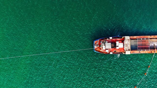 Red cargo ship with tow line viewed from directly above, Black Sea. Overhead drone shot of rust red barge hull against vivid green water, with steel cable extending diagonally across frame.