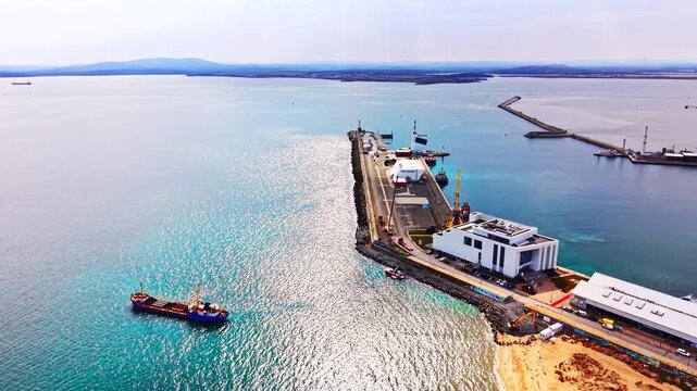 Aerial view of port peninsula with cargo ship and bay panorama. Narrow quay stretching into shimmering sea, white terminal building, loading crane, and distant mountain silhouette.