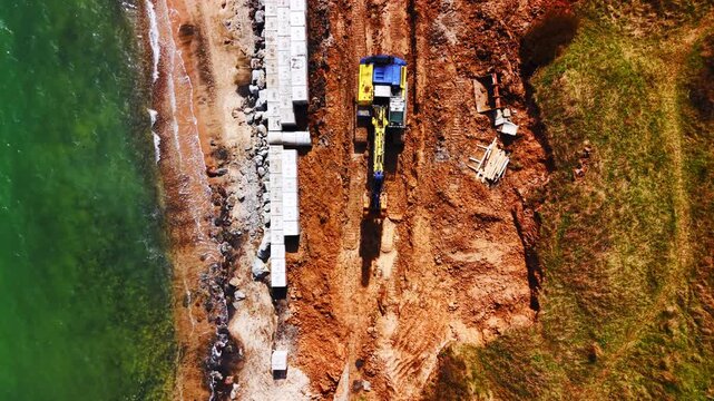 Overhead view of excavator placing concrete blocks on coastal seawall. Top-down drone view of yellow and blue excavator alongside row of precast concrete segment beside rocky shore and green sea.