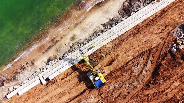 Seawall construction along coastline. Drone panorama shows excavator, mixer truck, precast concrete block and rebar beside turquoise sea and green farmland.