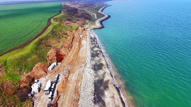 Excavator placing blocks on coastal seawall with sea and rock. Overhead shot of yellow and blue excavator working beside new precast wall, rocky riprap and green water at left.