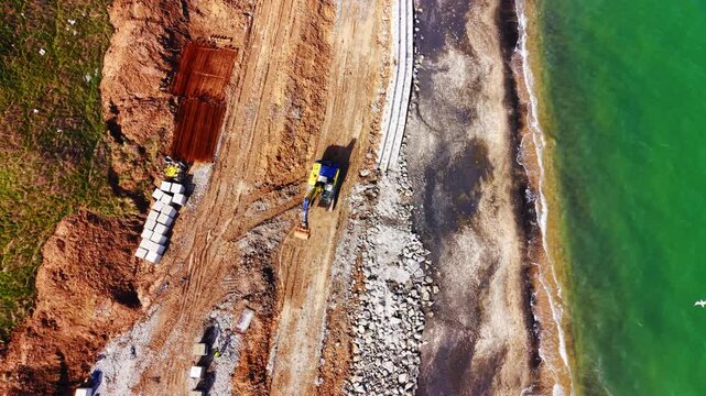 Top-down view of seawall construction with excavator and materials on coast. Excavator, concrete block row, rebar stack and sandy embankment beside green sea and rocky shore.