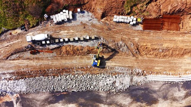 Coastal seawall block installation with two excavators. Aerial view of yellow and blue excavator beside precast concrete block row, rocky beach, riprap and green sea at edge.