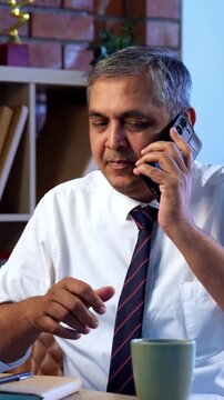 Indian executive working on computer while attending phone call at desk in modern office, Asian corporate professional multitasking with focus and communication skills in contemporary workplace setup