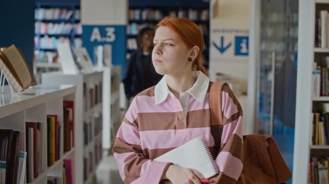 Tracking medium slow motion shot of Caucasian female teenager holding notebook walking in library while other students walk in background. For university enrollment promos and educational marketing