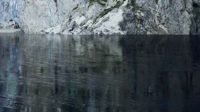 The scene shows clear water reflecting steep cliffs and rocks. The cliffs are rocky with patches of vegetation. The sun is shining on the water, creating a bright spot.