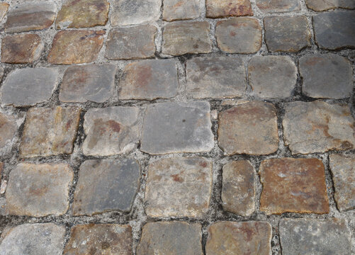 A fragment of an ancient cobblestone pavement made of massive stone blocks in various shades. The textured road surface shows signs of time, unevenness, and natural colors from gray to brown.