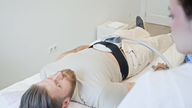 Young adult Caucasian man lying on clinic table during electromagnetic muscle stimulation treatment on abdomen. Ideal for promoting non-invasive body contouring services