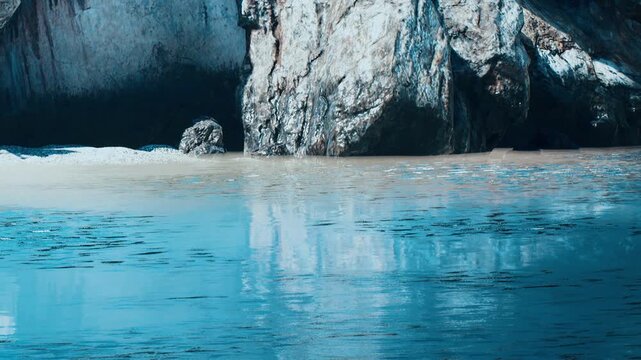 Water meets a rocky shore with clear reflections in the surface. The scene shows a natural area with ocean and cliffs. Sunlight creates a warm glow during the late afternoon hours.
