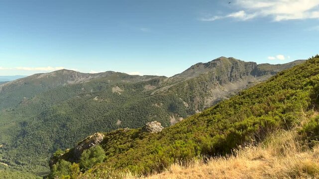 Panoramic mountain view with rolling hills and green valleys. Clear blue sky with scattered clouds above rugged terrain. Vegetation includes shrubs and grasses on sunlit slopes
