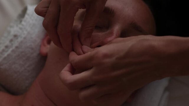 A professional close-up slow-motion video of a wellness facial massage treatment. A professional female masseuse gently and delicately massaging the glowing skin of a young woman's face at a luxury sp