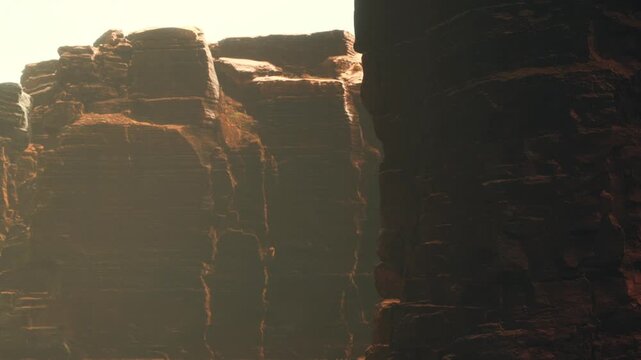 Tall rock formations rise in a canyon during the day. Sunlight shines on the cliffs creating shadows. The scene shows the rugged texture of the rocks.