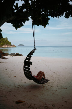 Person relaxes on swing under tree by beach in warm weather