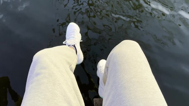 POV of male swinging legs on wood pier above calm lake. Summer sunlight reflects on water surface creating peaceful and immersive atmosphere. The scene conveys freedom and quiet escape. Slow motion
