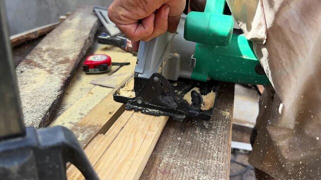 Arms of professional repairman sawing wooden construction with a circular saw at construction. Male hands of adult craftsman cuts wood using electric grind wheel at the site. Slow motion