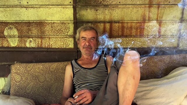 Portrait of homeless adult man sitting on worn couch placed and smoking outdoor. The setting underscores neglect and impermanence, presenting a raw and unfiltered glimpse of difficult living situation