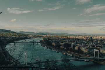Fototapeta premium ​Horizontal panoramic high angle view of a city river landscape with multiple historic bridges. Grand urban cityscape and mountain skyline under a cloudy teal sky, shot in Budapest, Hungary.