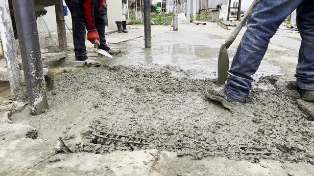 Male hands in protective gloves uses trowel to spread and level fresh cement in yard. Another worker distributes and moves the mixture with a shovel preparing the material during construction process