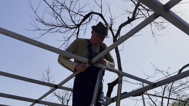 Male laborer uses a hammer to strike welded metal joints on carport frame. Adult worker refines and secures the structure, carefully adjusting welded connections metal installation work at the site