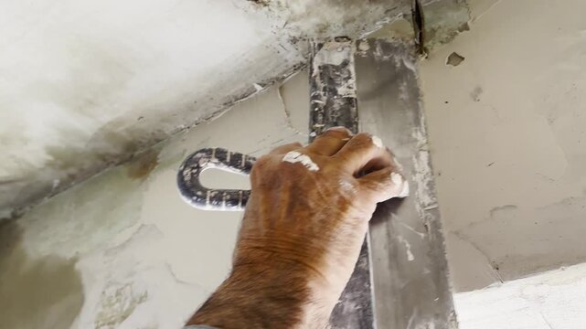 Male hand uses a putty knife to apply and spread fresh putty on interior wall. Worker carefully levels the material preparing surface for finishing and creating smooth base for further renovation work
