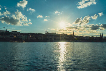 Fototapeta premium ​Horizontal scenic view of a city river skyline at sunset. Sunny urban landscape with a bright sun reflecting on calm water ripples under a cloudy sky, shot in Budapest, Hungary