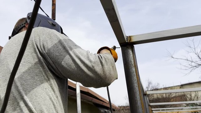 Manual laborer in protective mask weld iron detail on carport frame. Professional young repairman with welding machine working at construction. Male welder operating with metal at the site. Close up