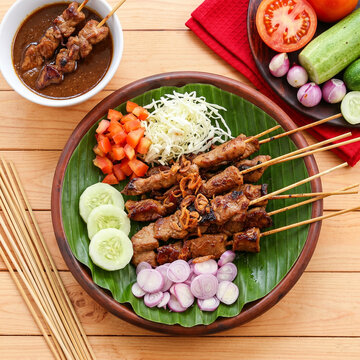 Indonesian Traditional Sate Kambing Mutton Satay on Clay Plate with Banana Leaf, Flat Lay View of Asian Skewered Grilled Meat with Peanut Sauce, Raw Shallots and Vegetables on Wooden Table
