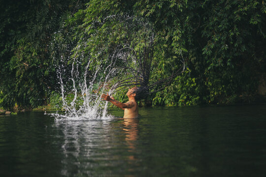 Man with long dreadlocks flipping hair back creating water splash arc in tropical river, ethnic guy bathing in nature rainforest jungle