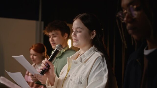 Side view of diverse teenagers rehearsing performance on stage, Caucasian teenage girl singing into microphone while holding script. For high school theater, arts education, and youth talent concepts