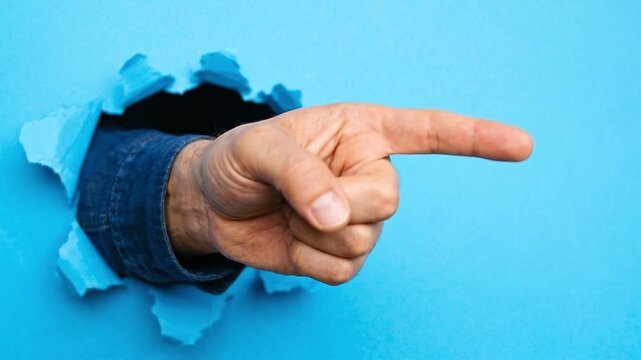 Tracking zoom close up of hand pointing through torn blue paper background, male arm extending from dark hole in studio, playful assertive directional gesture