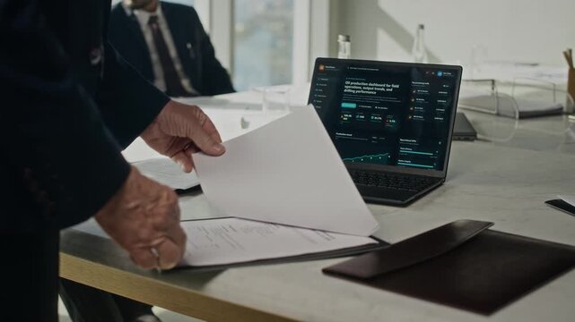 Cropped shot of businessman flipping through paper documents on clipboard next to laptop showing oil production dashboard. Footage is perfect for energy sector marketing videos
