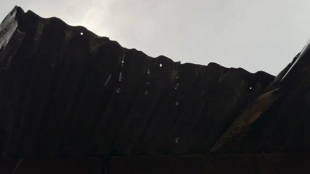 Rainwater drips from a damaged corrugated metal roof against a muted, overcast sky 