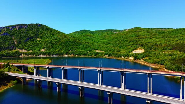 Bridge spans over water, Bulgaria. Side perspective of parallel railway and road bridges extending across deep reservoir flanked by wooded slopes.