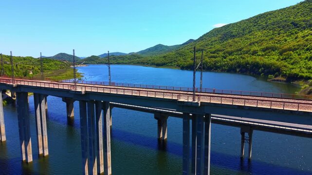 Railway bridge over reservoir. Elevated view of electrified track on concrete supports above blue water and hilly terrain.