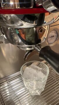 Pouring fresh espresso shot from a professional coffee machine into a glass with ice cubes