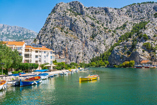 Picturesque view of small colorful boats moored on the Cetina river in Omis, Croatia, with massive karst mountains and white buildings under a clear blue sky in summer