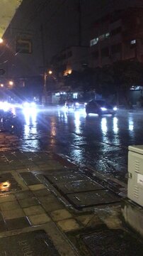 Cars driving in the rain at night in Bangkok Thailand viewed from the street side as they pass by with lights reflecting on the wet road surface. 