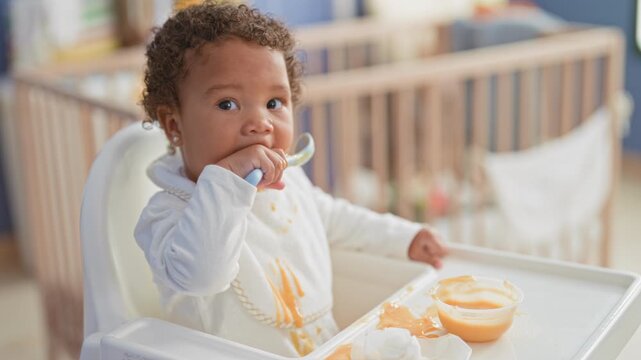 Little girl eating puree, holds spoon to mouth with bib stained and puree dripping from chin in building highchair; playful curiosity.