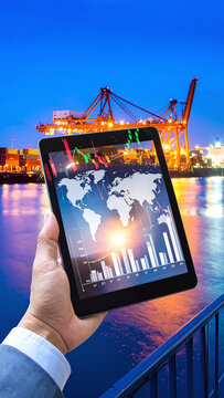 Businessman holding a tablet with global financial charts and a world map against a glowing cargo port at dusk, representing modern trade, digital logistics, and commerce.