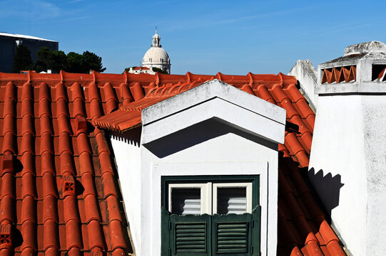 sloped red clay residential roof closeup. spanish or Portuguese style curved tile roofing. dormer with white and green wood window, old church cupola and tower background. travel and tourism concept