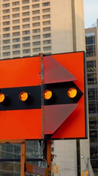Orange construction detour sign with arrow lights against urban high-rise buildings, flashing in alternating frames as signal bulbs shift from dark to amber, directing traffic through work zone.