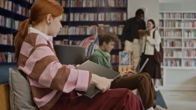 Side view shot of Caucasian teenage girl typing on laptop, closing lid, packing bag, and leaving library while other multiethnic teenagers study. Video for educational promotions or student life vlogs