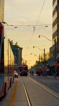 Streetcar travels along city tracks at sunset with pedestrians, overhead wires, and tall buildings lining roadway in warm evening light, urban transit passing through downtown street