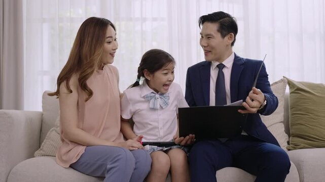 Happy Asian family, Mother, and Father sitting relaxing and looking at their daughter playing portable game console with joyful expression on sofa in living room at home. Dad in suit return from work.